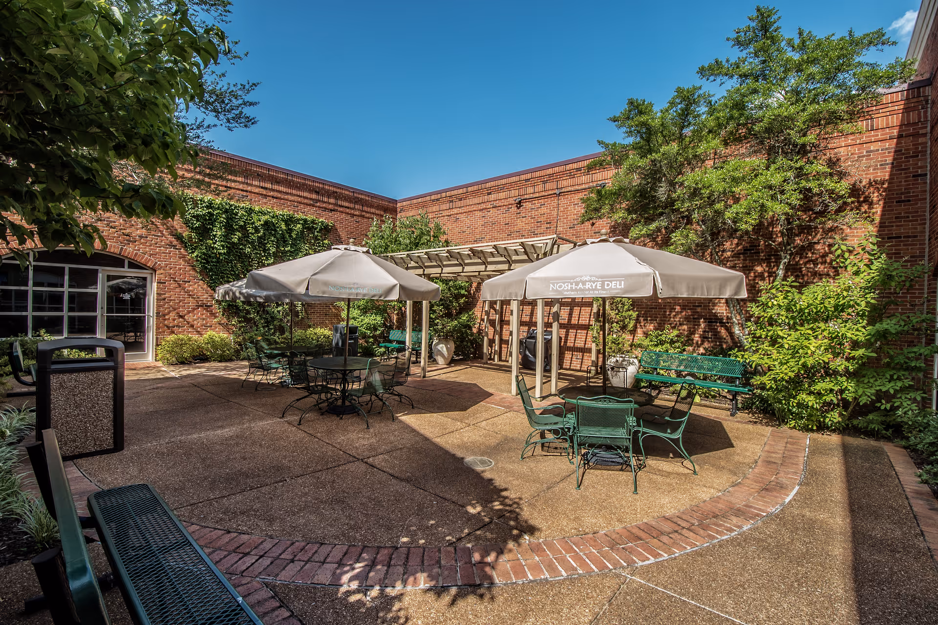 Sunny brick courtyard with patio tables, umbrellas, green metal chairs and benches under a clear blue sky.