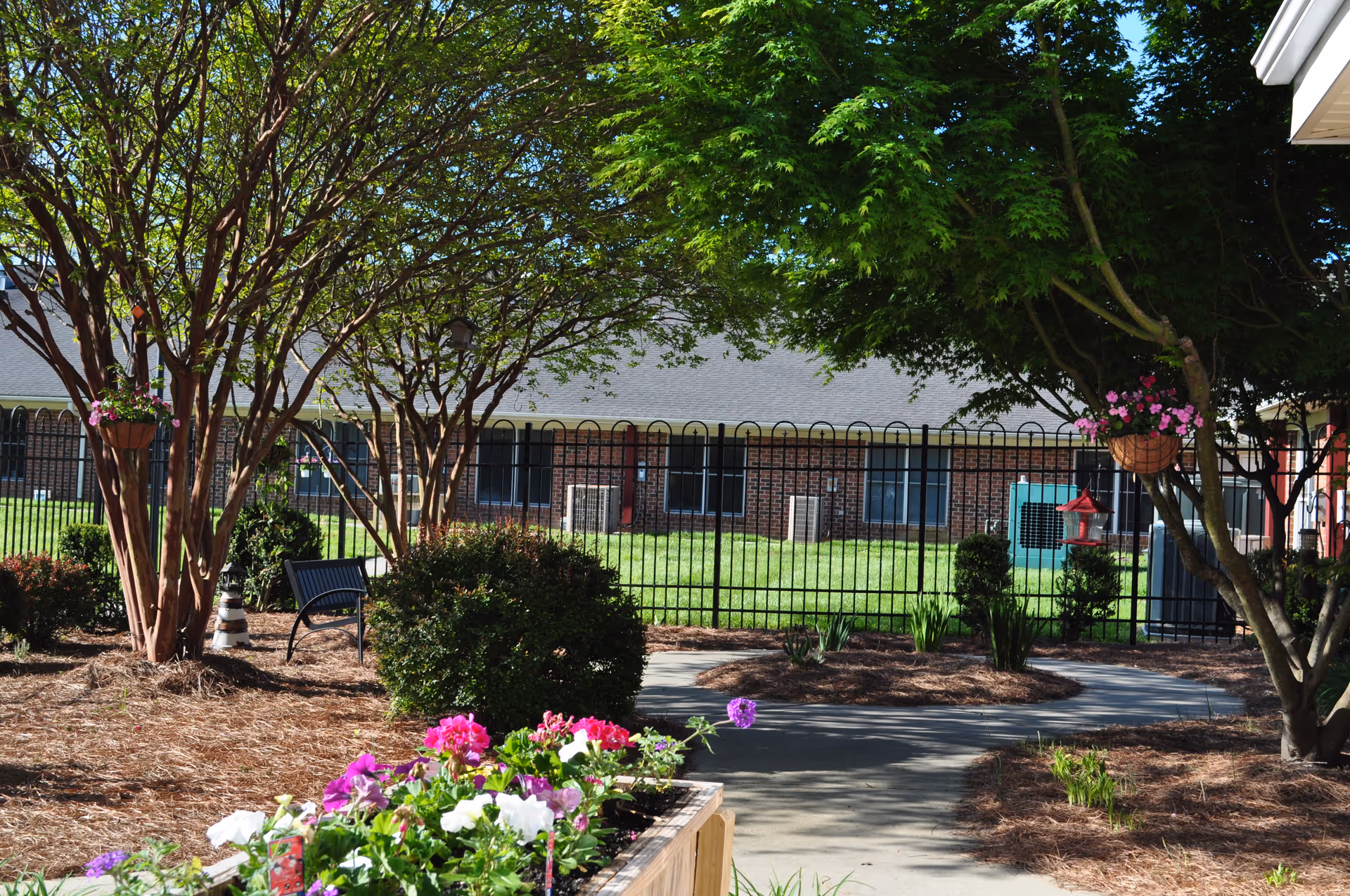 Landscaped courtyard with flowering planters, trees, a walkway and bench in front of a fenced brick building.