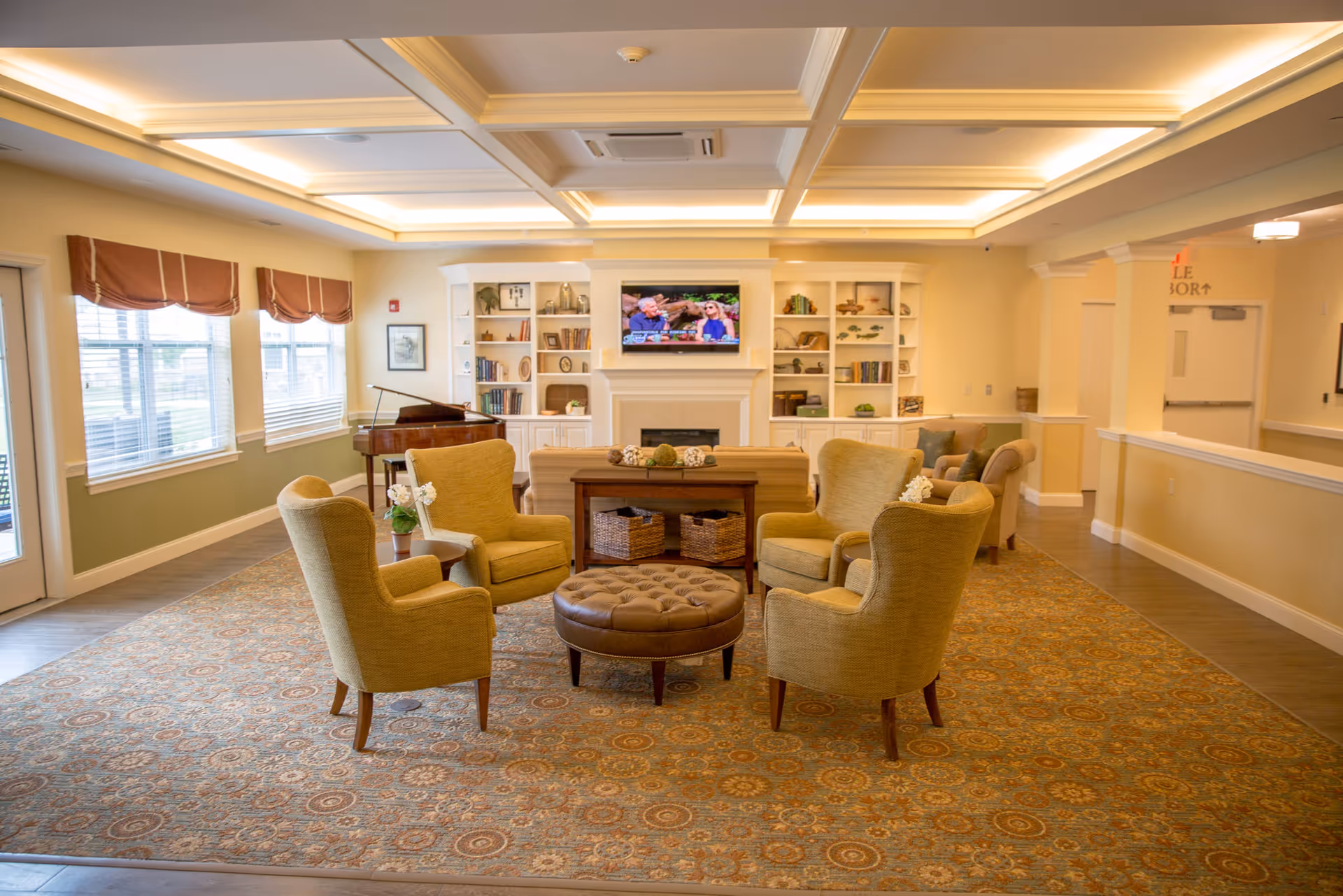 Well-lit senior living common room with four armchairs arranged around a central ottoman, a fireplace with TV and built-in bookshelves, and a piano by the windows.
