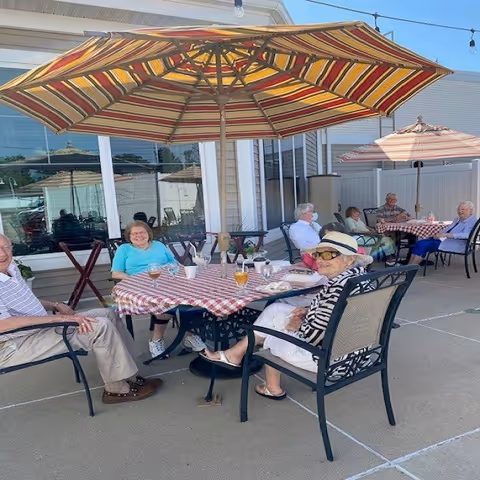 A group of elderly people sitting outdoors at tables with red and white checkered tablecloths under large striped umbrellas. They are enjoying drinks and socializing on a sunny day on a concrete patio next to a building with large windows.