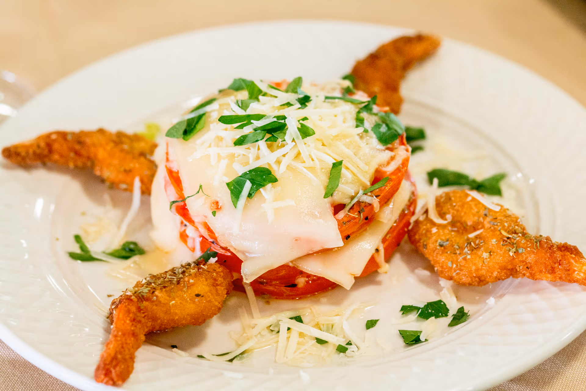 A plated dish featuring a stack of sliced tomatoes layered with melted cheese and topped with shredded cheese and chopped parsley, surrounded by four breaded and fried shrimp garnished with herbs on a white plate.