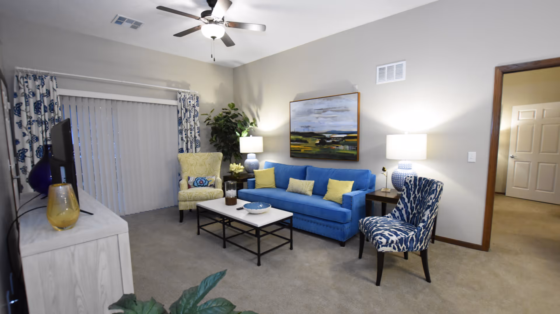 A cozy living room with a blue sofa adorned with yellow pillows, a patterned blue and white chair, and a yellow armchair. There is a white coffee table with decorative items in front of the sofa, a TV on a light-colored stand to the left, and two table lamps on either side of the sofa. A ceiling fan with a light is mounted on the ceiling, and a large painting hangs on the wall behind the sofa. Sliding glass doors with vertical blinds and patterned curtains are visible on the left side, and an open doorway leads to another room on the right.