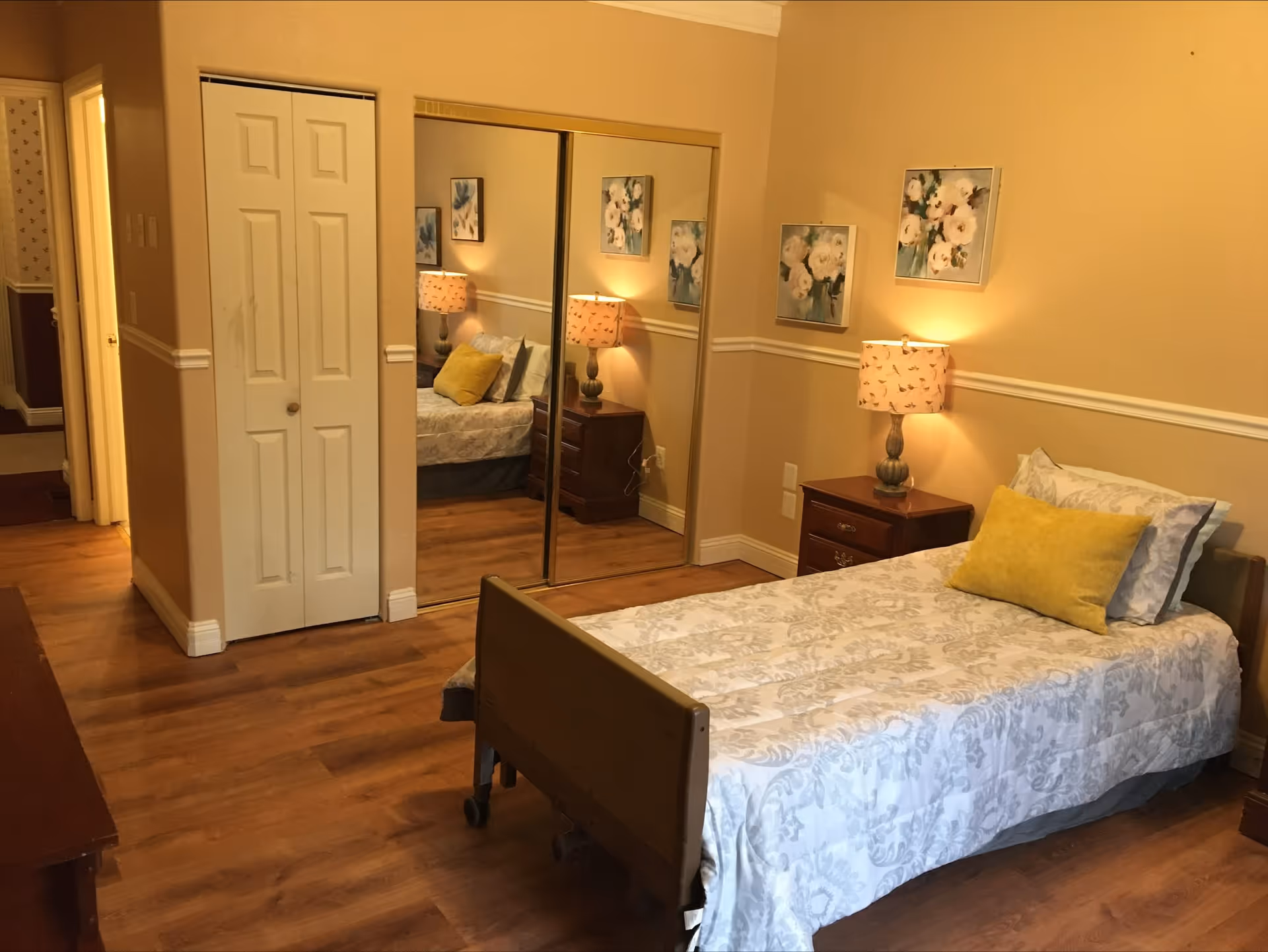 A cozy bedroom with a single bed featuring a white and gray patterned bedspread and a yellow accent pillow. There is a wooden nightstand with a decorative lamp and floral artwork on the beige walls. A closet with white double doors and mirrored sliding doors is visible, reflecting part of the room. The floor is wooden, and the room has warm lighting.