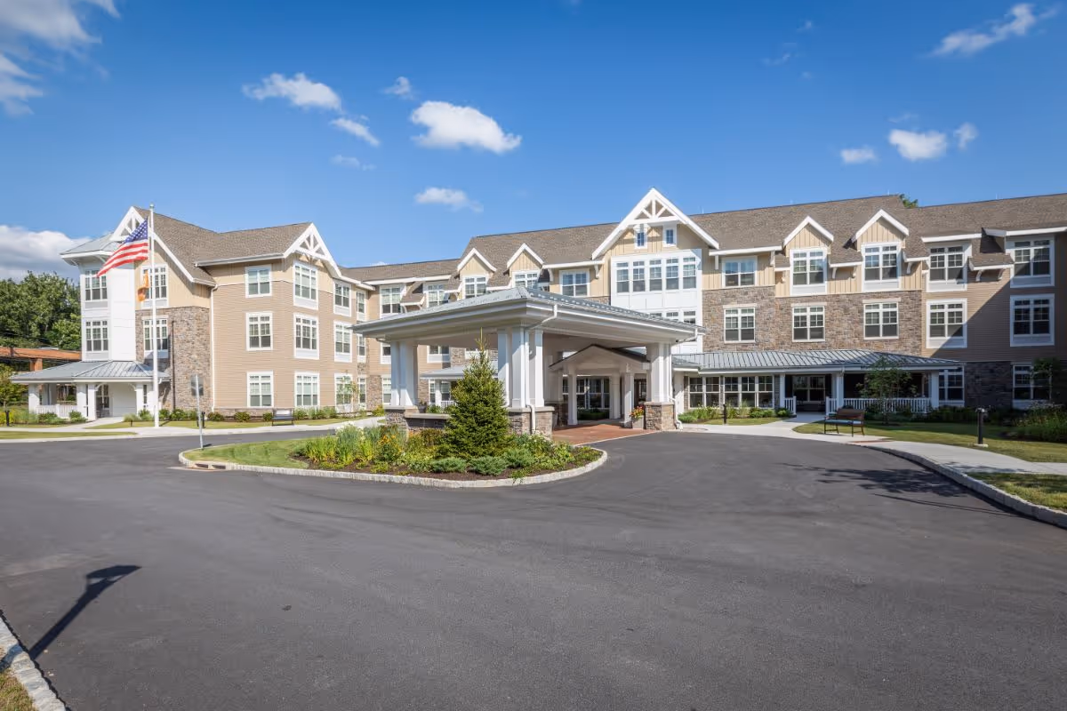 Front exterior of a senior living building with a covered porte-cochère, circular driveway, landscaped island, and an American flag under a blue sky.