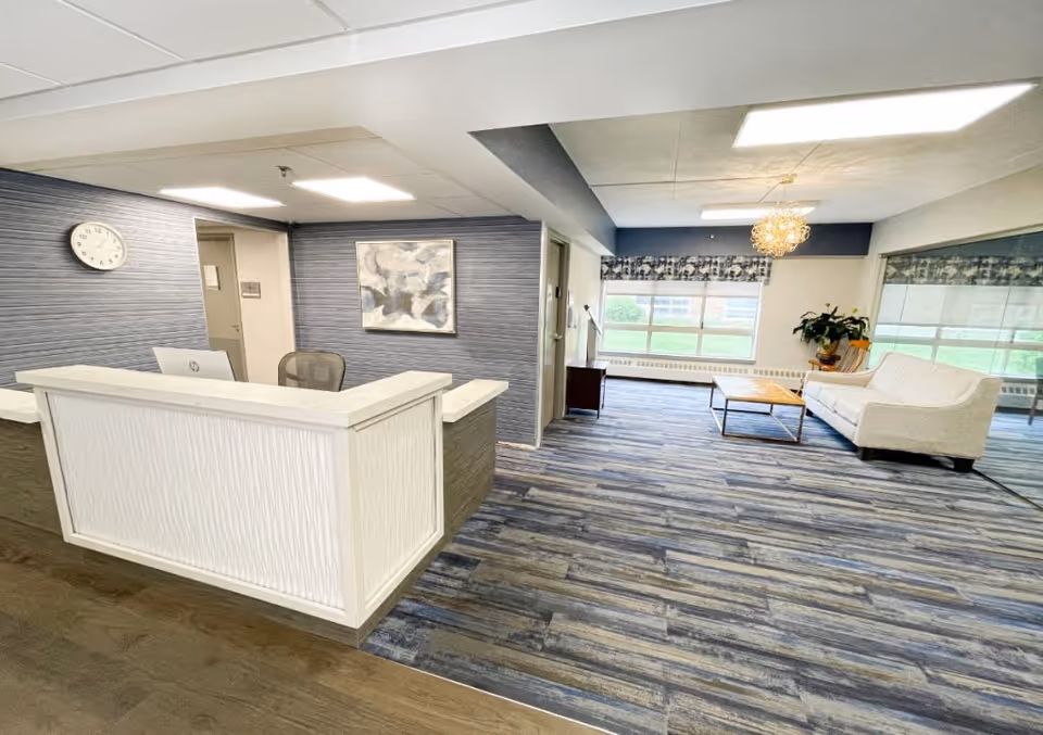 Reception area with a white front desk and office chair, a wall clock, abstract artwork on the wall, and a seating area with a white couch, wooden coffee table, and large windows letting in natural light.