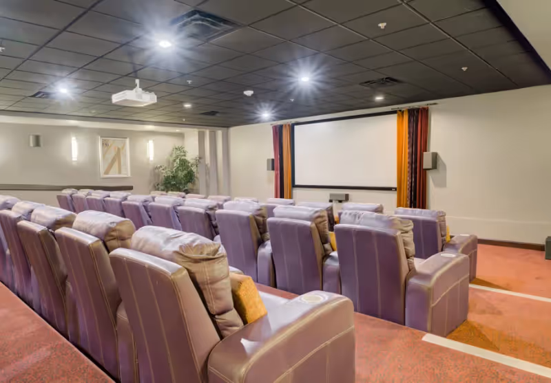 A cozy movie theater room with multiple rows of plush, purple reclining seats facing a large blank projection screen. The room has a black ceiling with recessed lighting, a projector mounted on the ceiling, and walls decorated with framed artwork and curtains on either side of the screen.