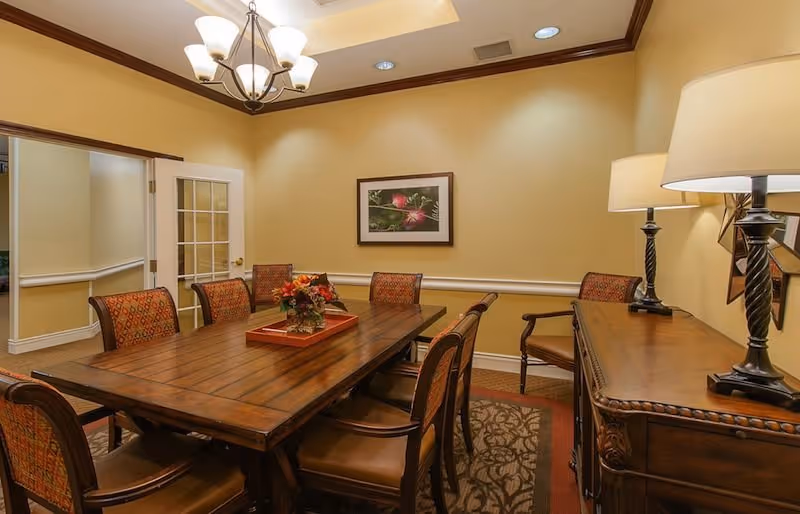A warmly lit dining room with a wooden rectangular table surrounded by eight upholstered chairs. The table has a floral centerpiece on a red tray. The room features beige walls, a framed floral picture, a chandelier overhead, and a wooden sideboard with two lamps on the right side. There is a door with glass panes leading to another room.