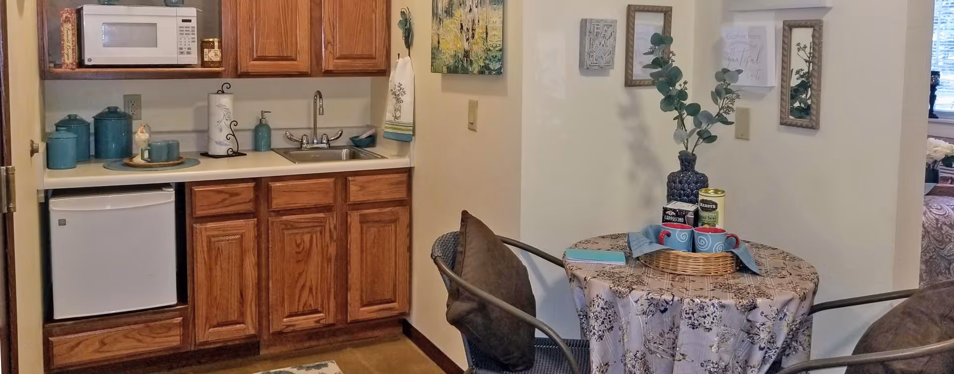 A small kitchenette area with wooden cabinets, a white microwave, a mini refrigerator, and a sink. To the right, there is a round table covered with a floral tablecloth, two chairs with cushions, and a decorative centerpiece with a vase and mugs. The walls have framed artwork and a towel hanging near the sink.
