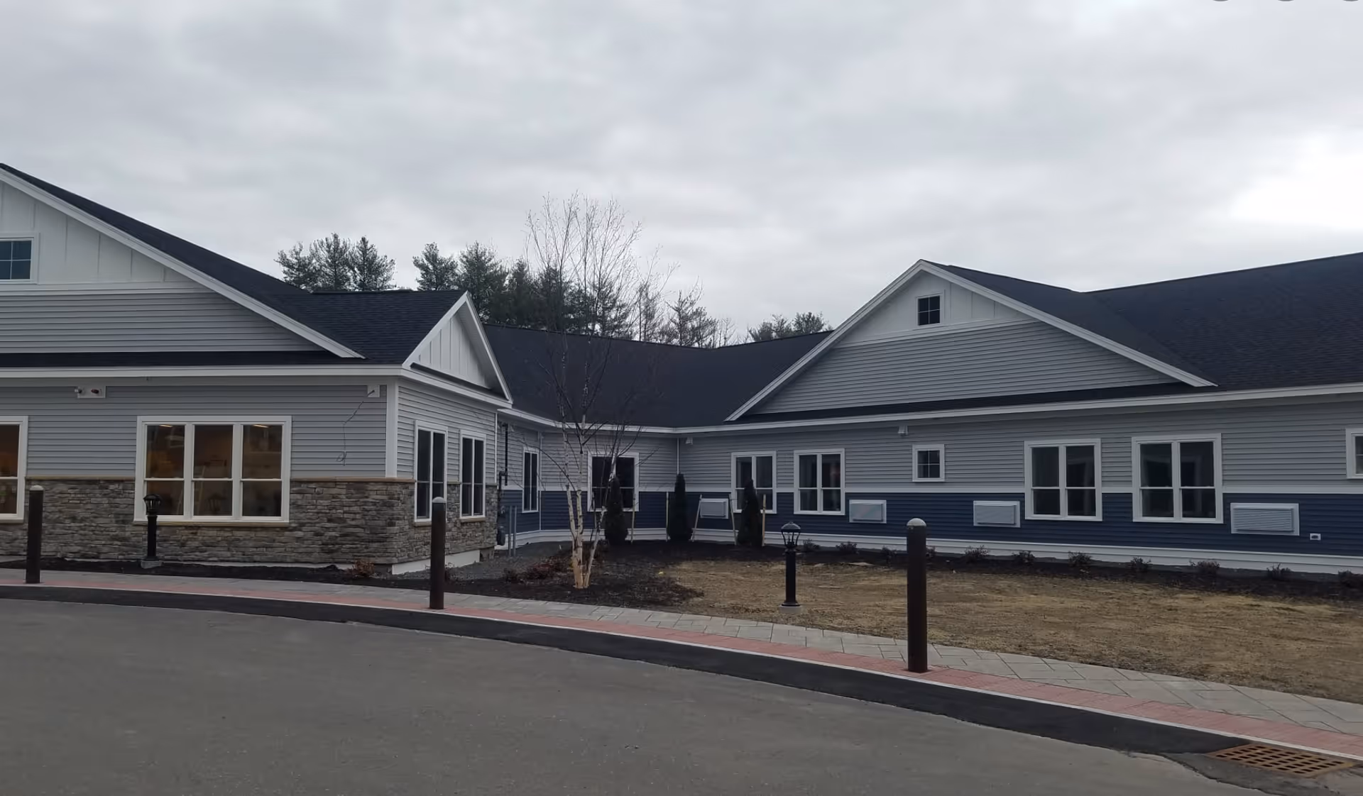 Exterior front of a single-story gray and blue senior living building with multiple windows, a driveway and overcast sky.