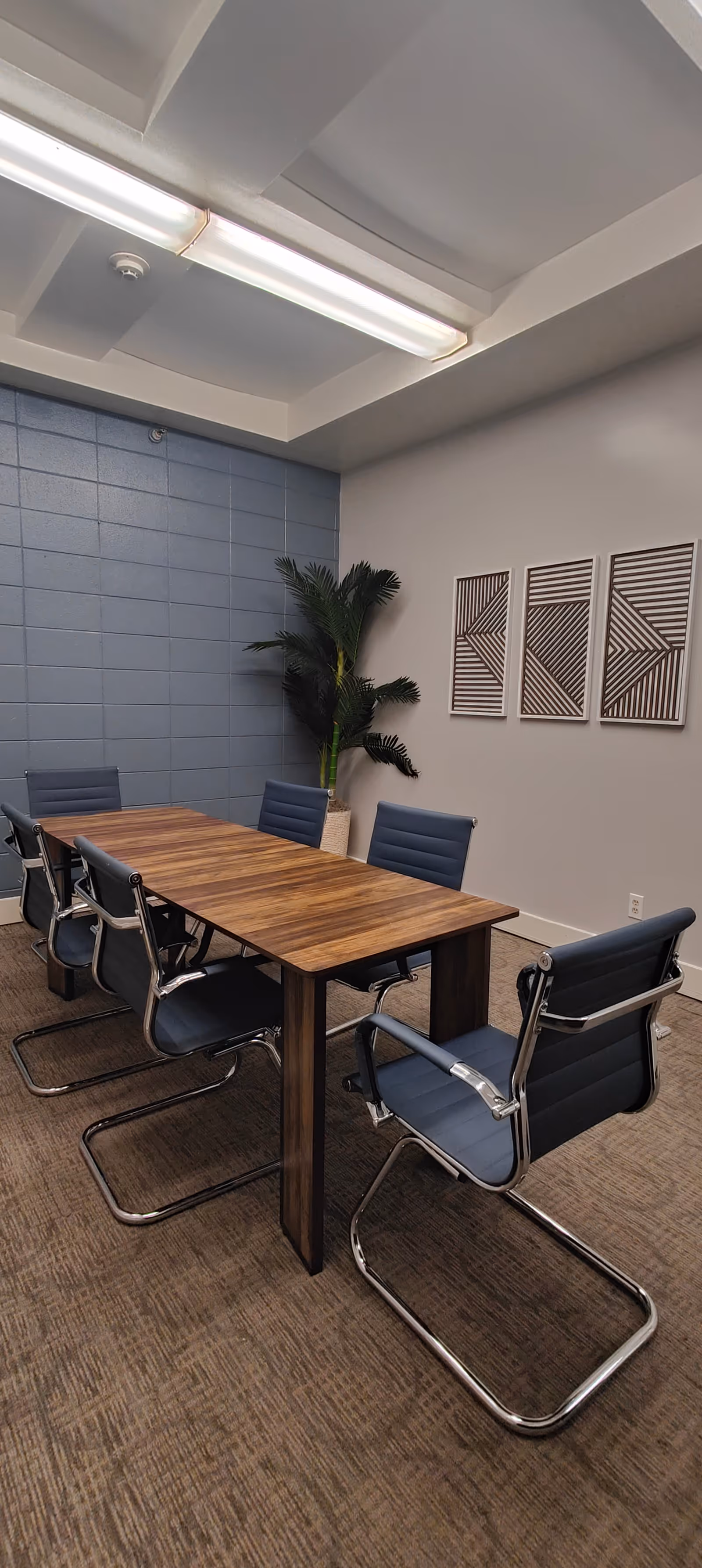 A modern conference room with a wooden rectangular table surrounded by six blue cushioned chairs with metal frames. The room has a blue accent wall with a potted plant in the corner and three framed geometric art pieces on a light gray wall. The ceiling features a recessed light fixture.