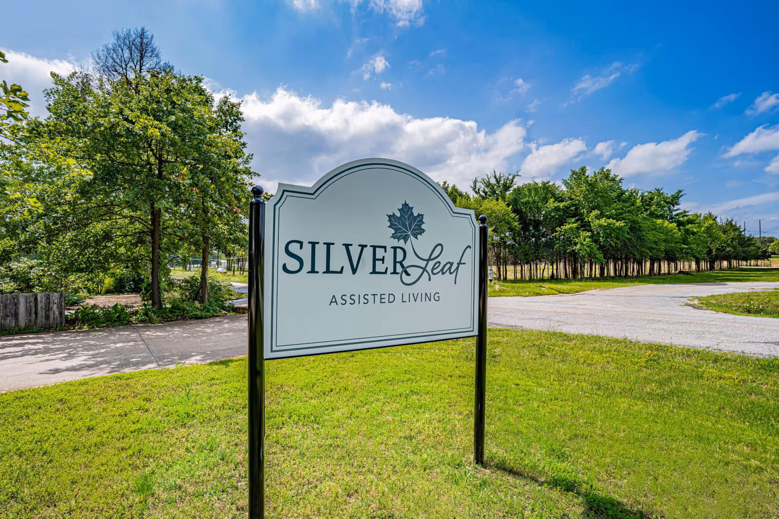 Outdoor view of a Silver Leaf Assisted Living sign on a grassy area with trees and a partly cloudy blue sky in the background.