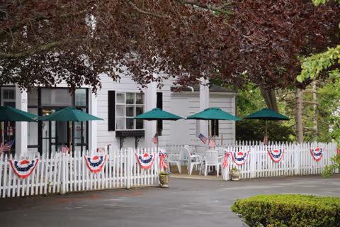 White-sided building front with a picket-fenced patio decorated with red, white, and blue bunting and small American flags, green umbrellas and white outdoor chairs under leafy trees.