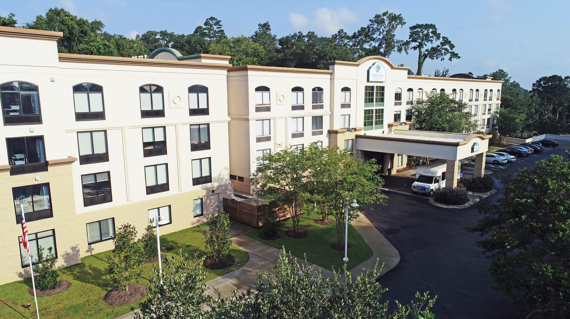 Exterior view of a multi-story senior living facility building with a covered entrance and parking lot, surrounded by trees and greenery under a clear sky.