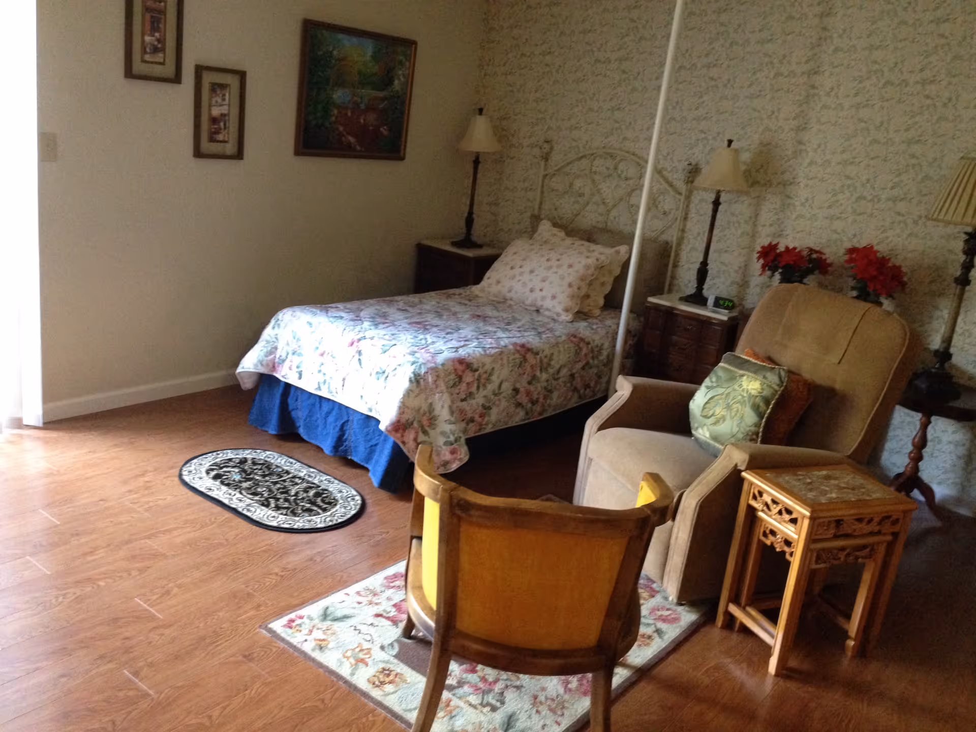 A cozy bedroom with a single bed covered in floral bedding, a wooden floor, a small oval rug, and two chairs with a small wooden side table. The walls have framed pictures and a floral wallpaper. There are two lamps on bedside tables and a few red flowers on one of the tables.