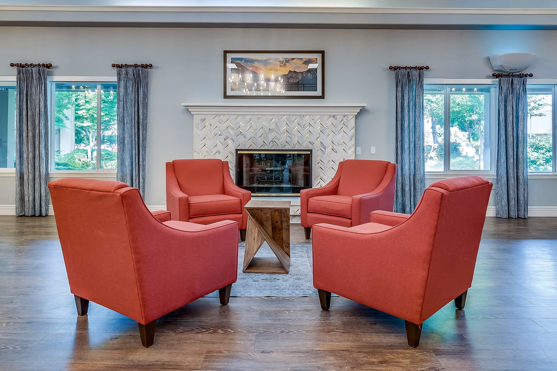 A cozy seating area with four red armchairs arranged around a small wooden coffee table in front of a decorative fireplace. The room has large windows with blue curtains, allowing natural light to brighten the space. A framed landscape painting hangs above the fireplace.