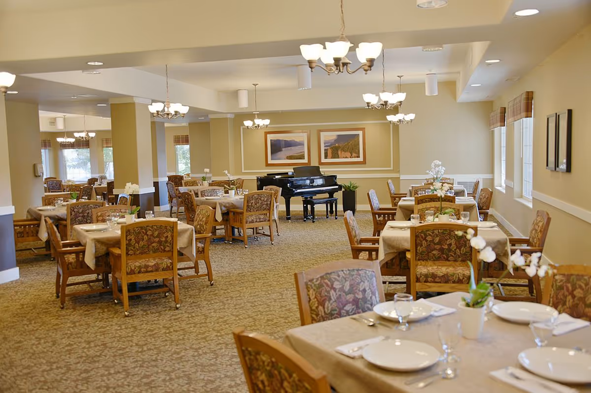 A spacious dining room with multiple tables covered in beige tablecloths, each set with white plates, glasses, and silverware. The chairs have floral upholstery. The room is well-lit with chandeliers hanging from the ceiling. At the far end of the room, there is a black grand piano and two framed landscape paintings on the wall. Windows on the right side let in natural light.