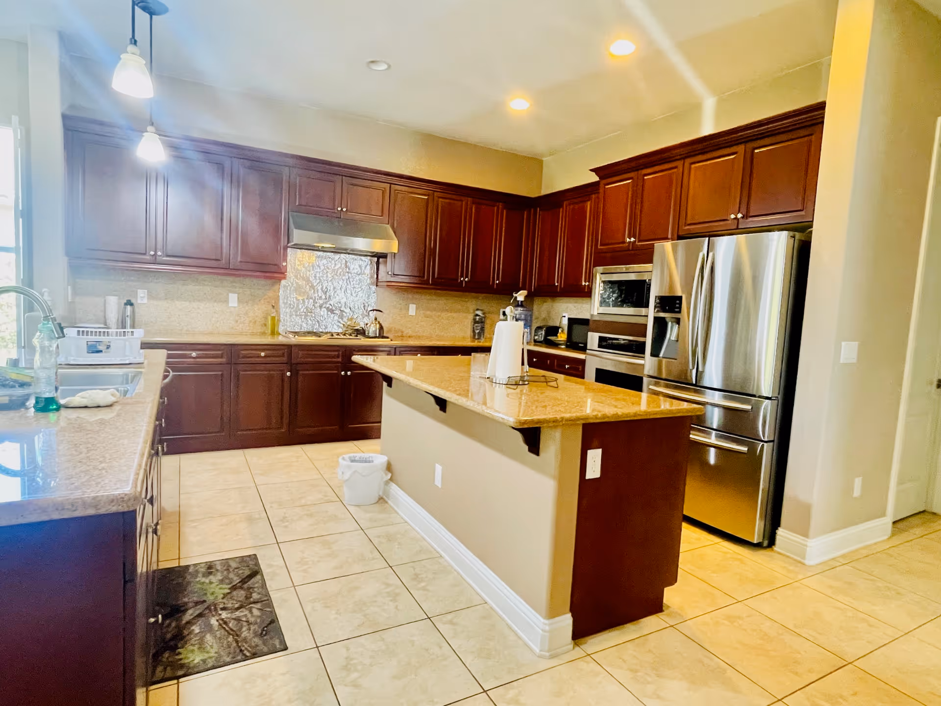Bright kitchen with dark wood cabinets, a large island with a granite countertop, stainless steel refrigerator, built-in microwave and oven, tiled floor, and pendant lights hanging from the ceiling.