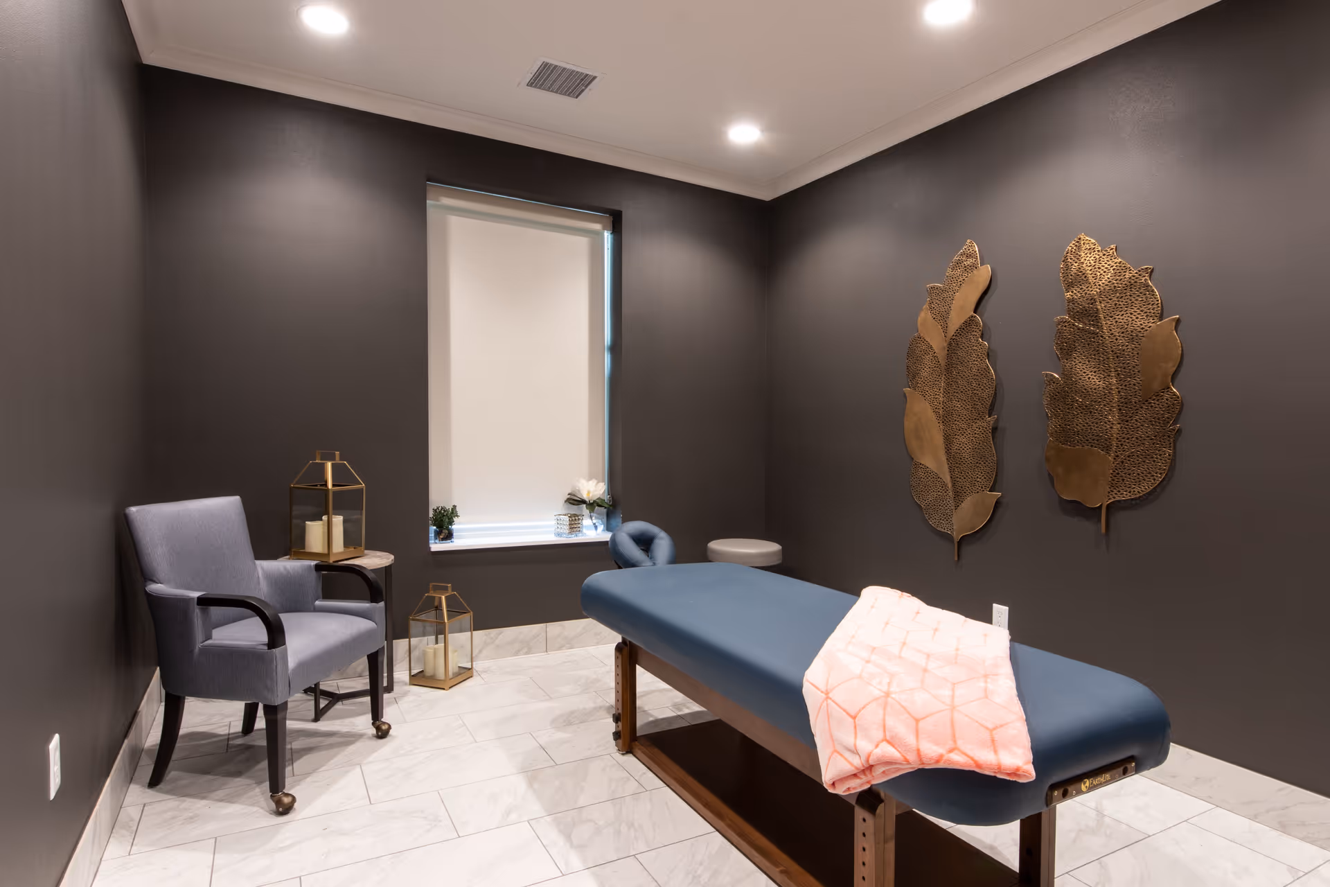 A calm treatment room with a massage table covered by a pink blanket, an upholstered chair, decorative metal leaf wall art, and soft recessed lighting.