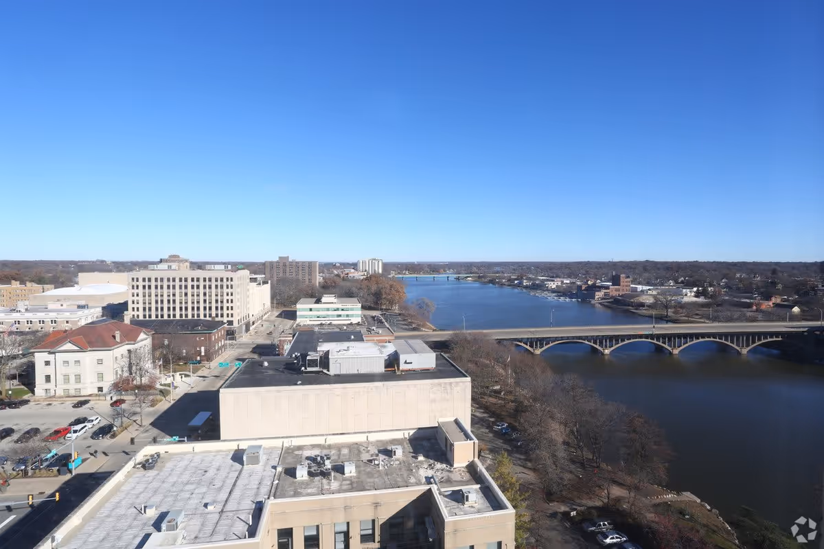 Aerial view of a cityscape featuring several buildings, a river with a bridge crossing it, and a clear blue sky.