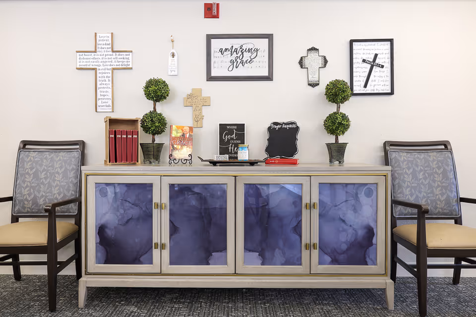 A decorated interior wall with a light-colored cabinet featuring four doors with purple abstract designs. On top of the cabinet are two small potted plants, several decorative crosses, framed inspirational quotes, books, and a small chalkboard labeled 'Prayer Requests.' Two cushioned chairs with patterned backs flank the cabinet on either side.