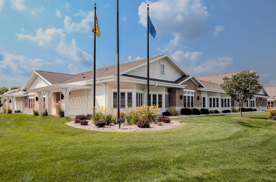 Exterior view of Countryside Manor, a single-story building with white siding and brick accents, surrounded by a well-maintained lawn, flower beds, and three flagpoles with flags against a partly cloudy sky.