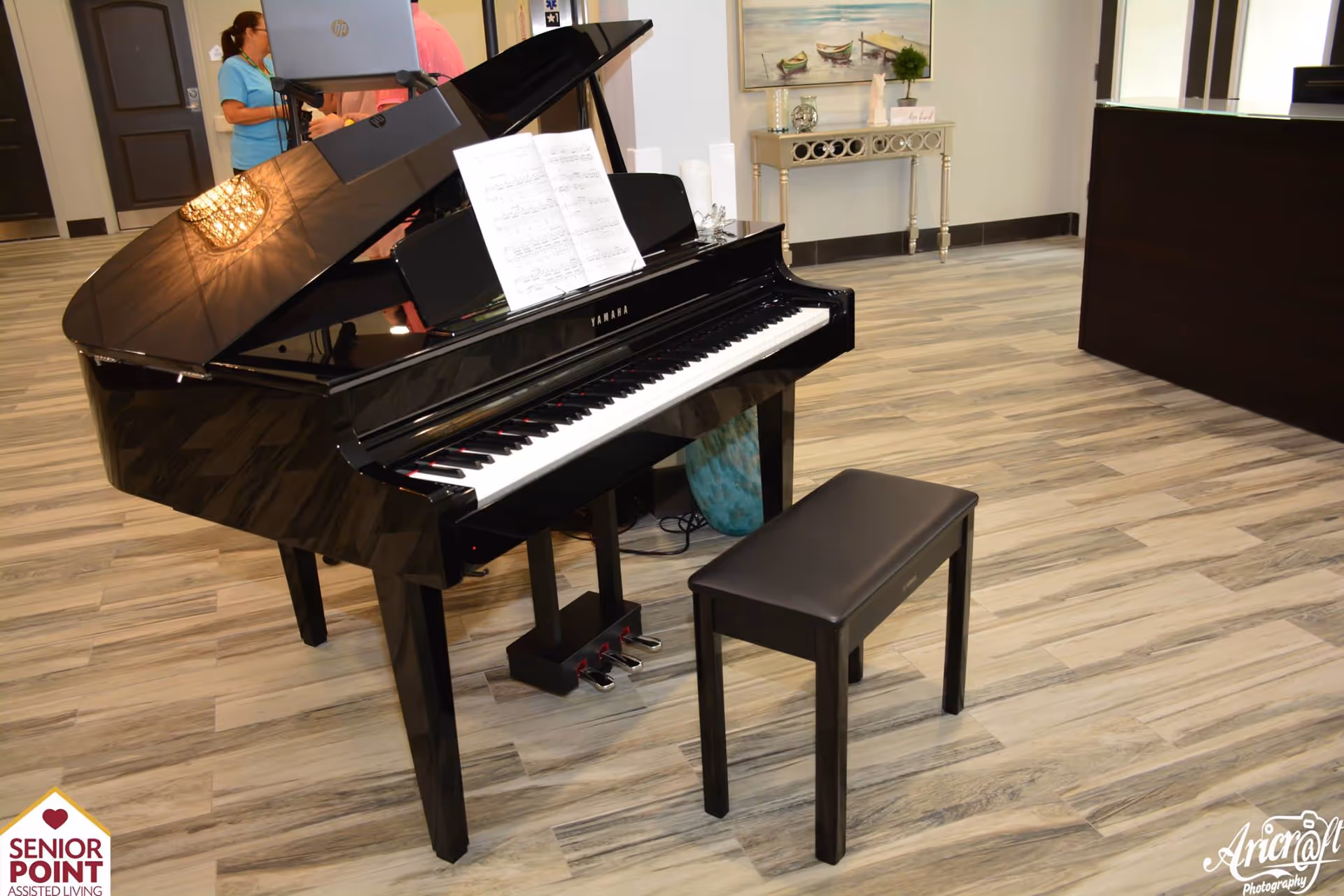A black Yamaha grand piano with an open lid and sheet music on the stand, accompanied by a black piano bench, situated in a room with light wood flooring. In the background, there is a decorative console table with a painting of boats on the wall above it, and part of a reception desk is visible. Two people are partially visible near the door in the background.