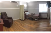 A rehabilitation and nursing facility room with a hospital bed, a brown recliner chair, a small table with a tray, a window with blinds, and a wall-mounted television. The room has wood flooring and light gray walls with a white chair rail.