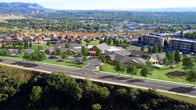 Aerial view of a residential area featuring a cluster of single-story buildings surrounded by greenery and trees, with a multi-story building in the background and a road running in the foreground. Mountains and a sprawling cityscape are visible in the distance under a clear sky.