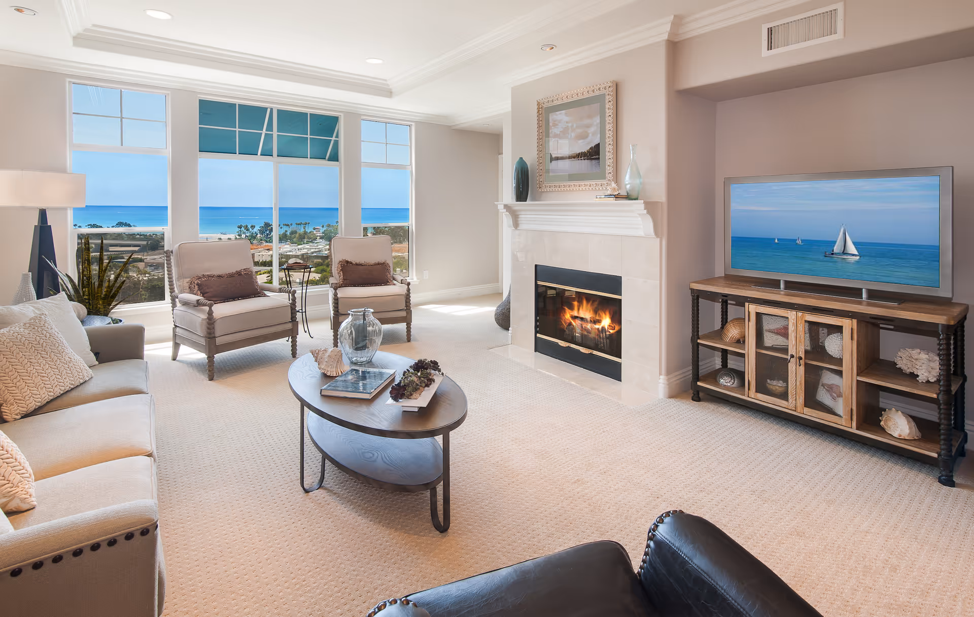 Bright living room with large windows offering an ocean view. The room features a beige carpet, a fireplace with a fire burning, a flat-screen TV on a wooden stand, a beige sofa with cushions, two beige armchairs, a black leather chair, and a coffee table with decorative items.