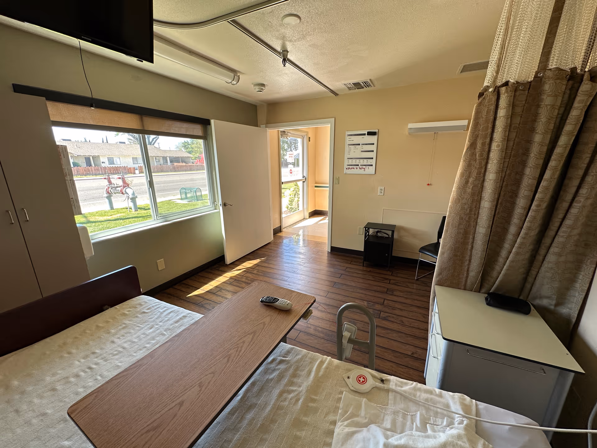 A nursing facility patient room with a bed, overbed table, window, open door, TV, and a privacy curtain.