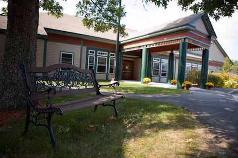 Exterior view of a senior living center with a decorative bench, path, and potted mums.
