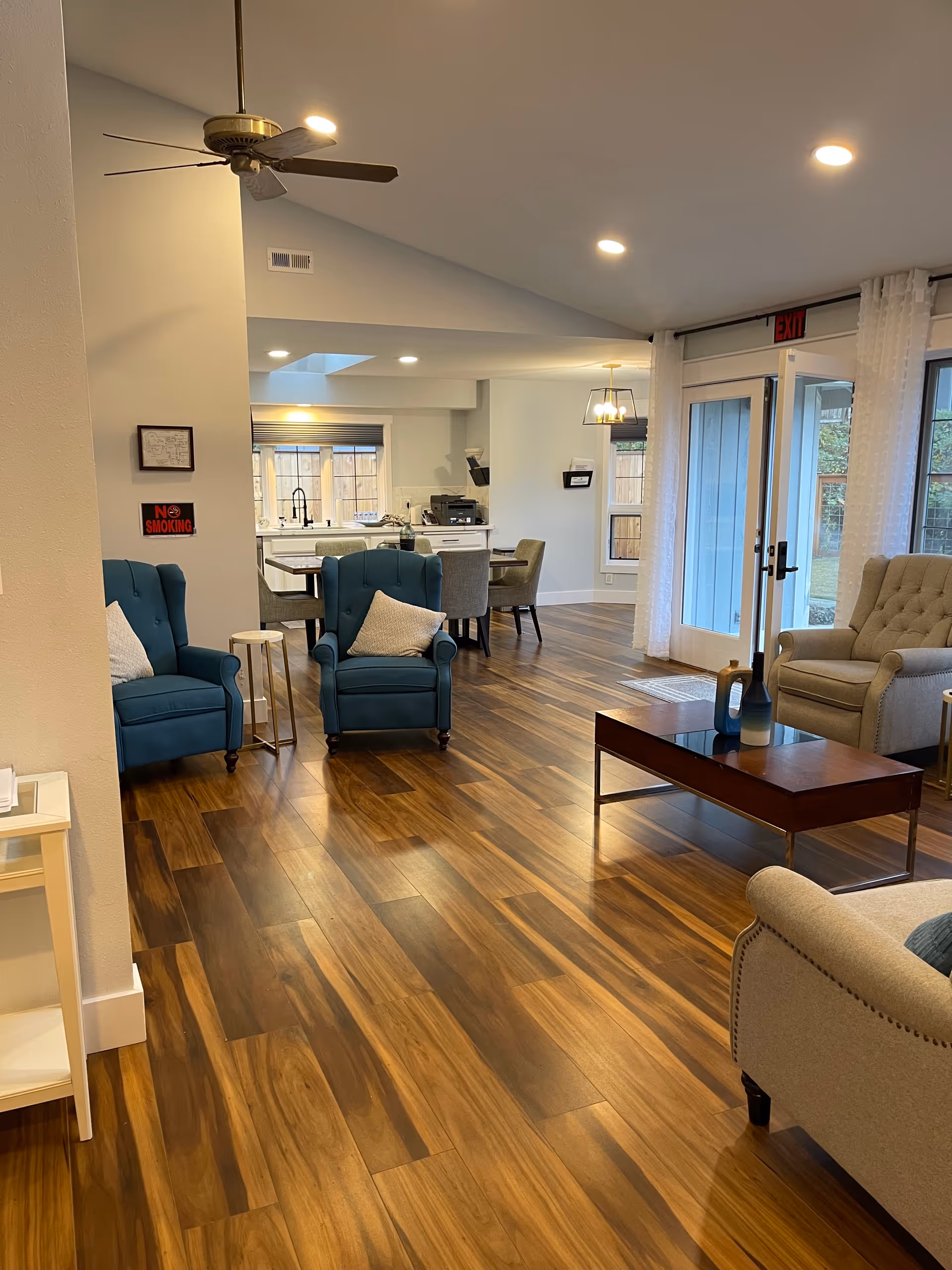 A spacious and well-lit living area with wooden flooring, featuring two blue armchairs with white pillows, a beige armchair, a wooden coffee table with decorative items, and a dining area with a table and chairs in the background. Large windows and glass doors with sheer white curtains allow natural light to enter the room. A ceiling fan and recessed lighting are visible on the ceiling.