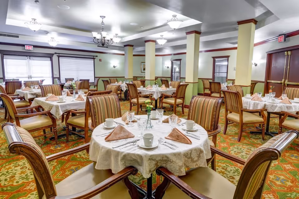Formal dining room with round tables set with napkins, dishes and glassware, surrounded by upholstered chairs and decorative lighting.