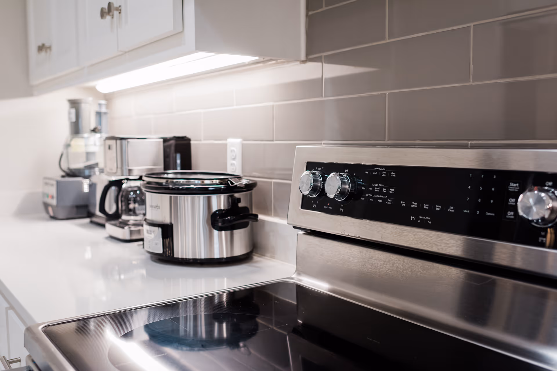 Close-up view of a modern kitchen countertop featuring a stainless steel electric stove with control knobs, a slow cooker, a coffee maker, and a food processor against a gray tiled backsplash and white cabinets.