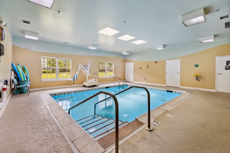 Indoor swimming pool with handrails and steps leading into the water, surrounded by beige walls and windows showing an outdoor view. Pool equipment and flotation devices are visible on the left side, and several closed doors are on the right wall under a light blue ceiling with multiple light fixtures.