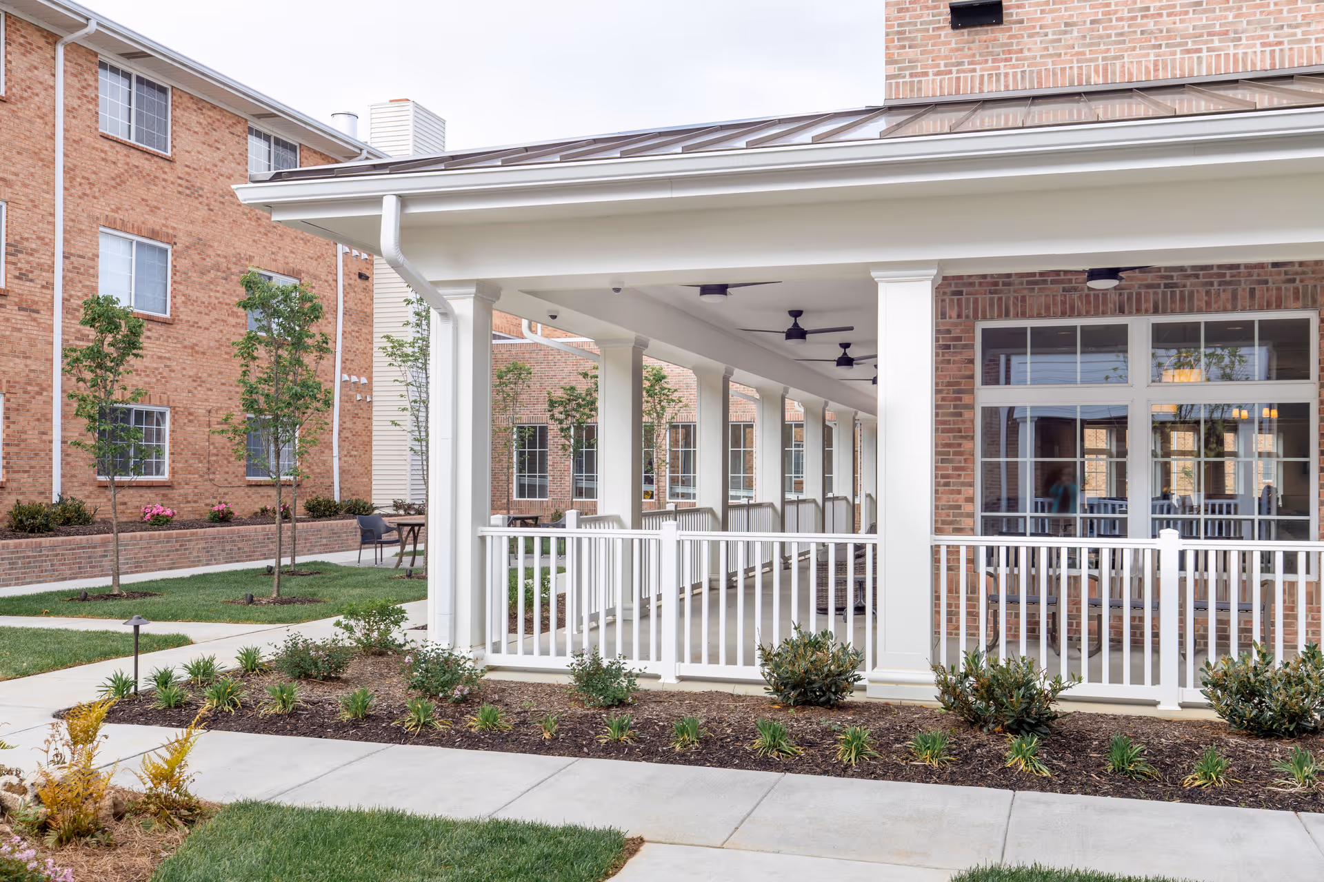 Outdoor covered walkway with white railings and columns at Friendship Towers, surrounded by landscaped garden beds and grass, with brick buildings in the background.