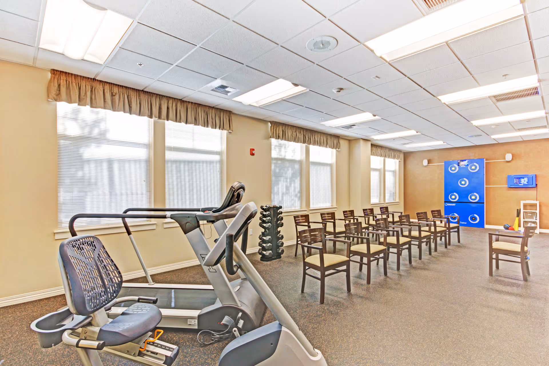A bright exercise room with large windows covered by beige valances. The room contains a treadmill, a recumbent exercise bike, a rack of dumbbells, and several wooden chairs arranged in rows facing a blue target board and a digital timer on the wall.