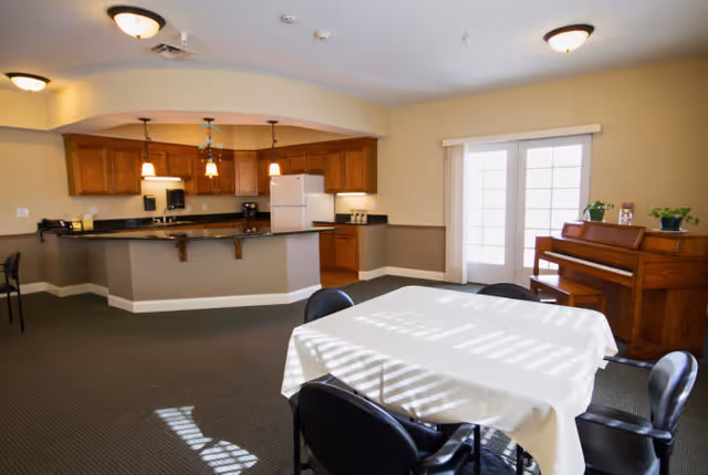 A spacious room with a kitchen area featuring wooden cabinets, a refrigerator, and a countertop with hanging pendant lights. In the foreground, there is a table covered with a white tablecloth surrounded by black chairs. To the right, there is a wooden piano with small plants on top, and a set of glass doors letting in natural light.