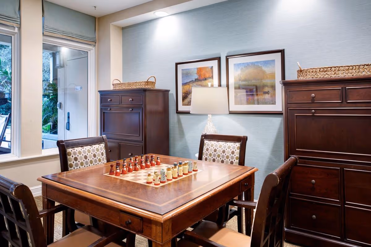 Cozy interior room with a wooden game table set up for chess, surrounded by chairs, lamps, framed art, and dark wood cabinets.