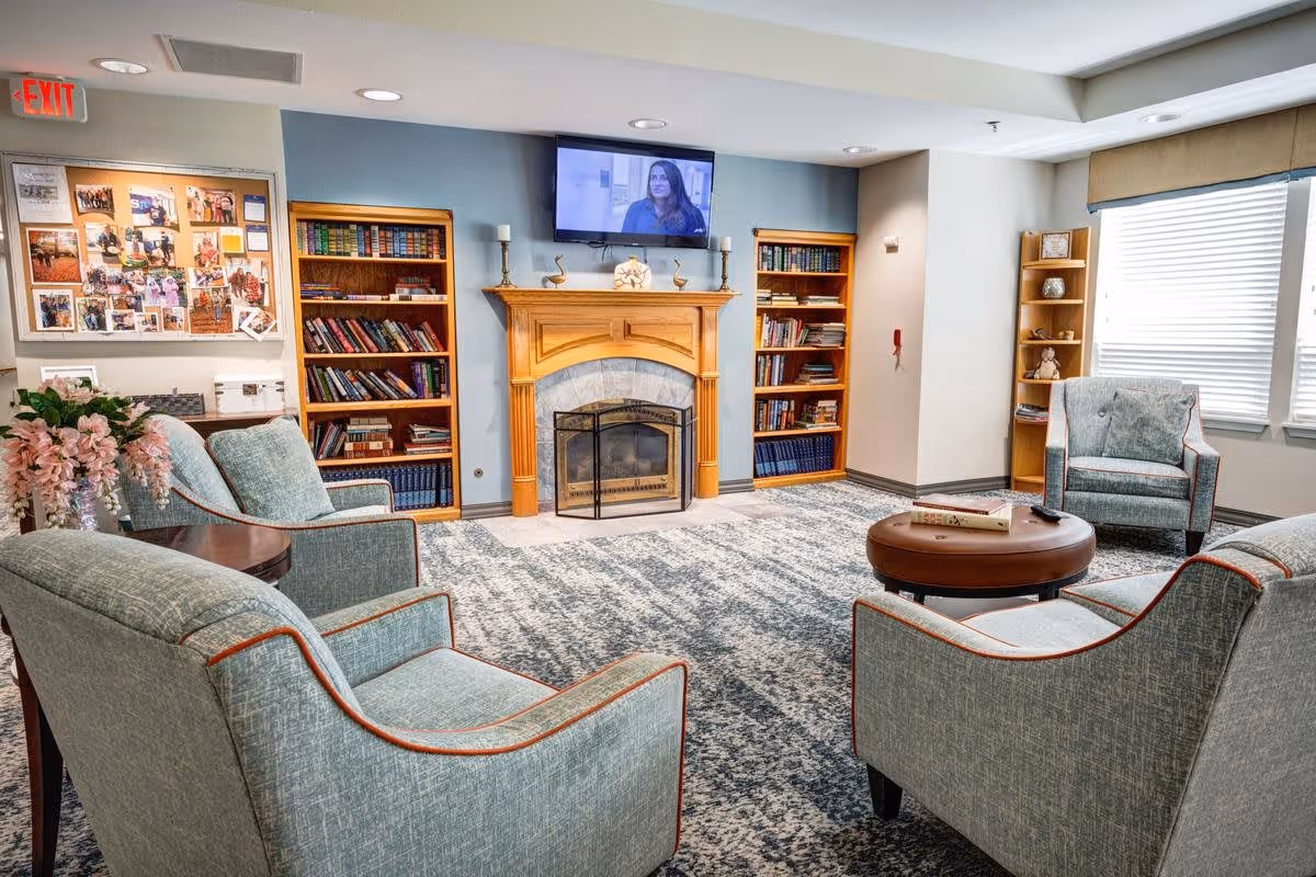 A cozy living room area with four upholstered armchairs arranged around a round wooden coffee table. The room features a fireplace with a wooden mantle, flanked by two bookshelves filled with books. Above the fireplace is a wall-mounted flat screen TV. There is a bulletin board with photos and notices on the left wall, and a window with blinds on the right side letting in natural light. The room has a patterned carpet and soft, neutral wall colors.