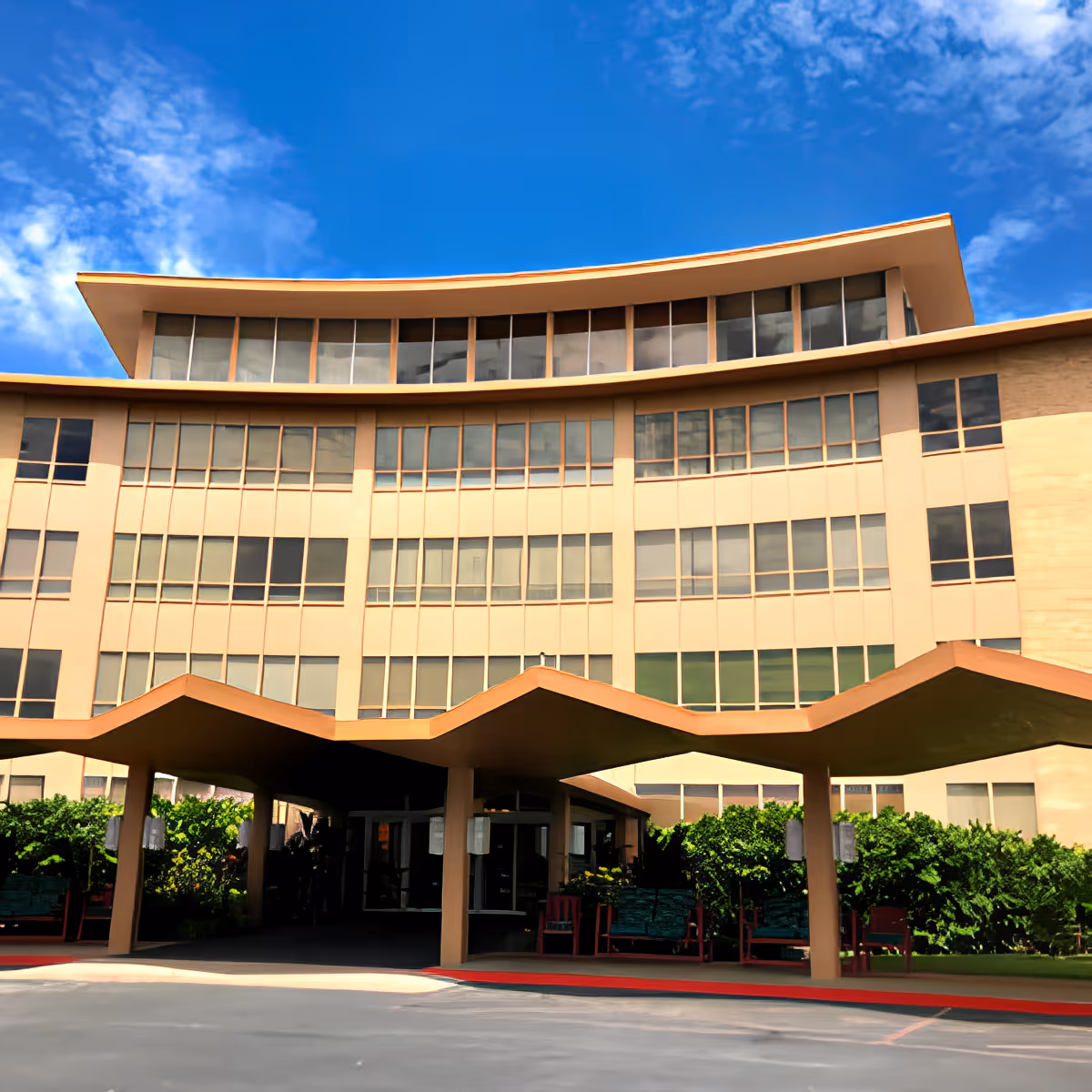 Exterior view of a multi-story senior living facility building with large windows and a distinctive wavy roof over the entrance. The sky is clear with some clouds, and there are green bushes and benches near the entrance.