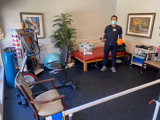 A small physical therapy or exercise room with various equipment including a stationary recumbent bike, exercise balls, dumbbells, and a padded treatment table. A person wearing a mask and holding an orange ball and a green dumbbell stands near the treatment table. The room has framed artwork on the walls and a potted plant in the corner.