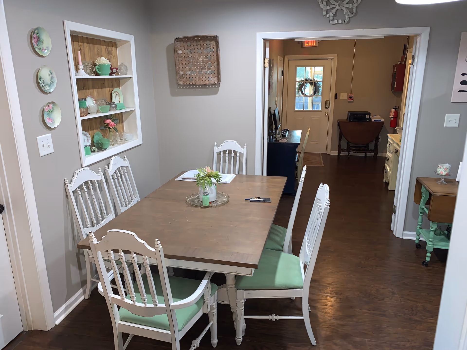 A dining room with a wooden table and six white chairs with green cushions. The walls are light gray with decorative plates and a shelf displaying glassware and small decorative items. There is a doorway leading to a hallway with a door at the end, and the floor is dark wood.