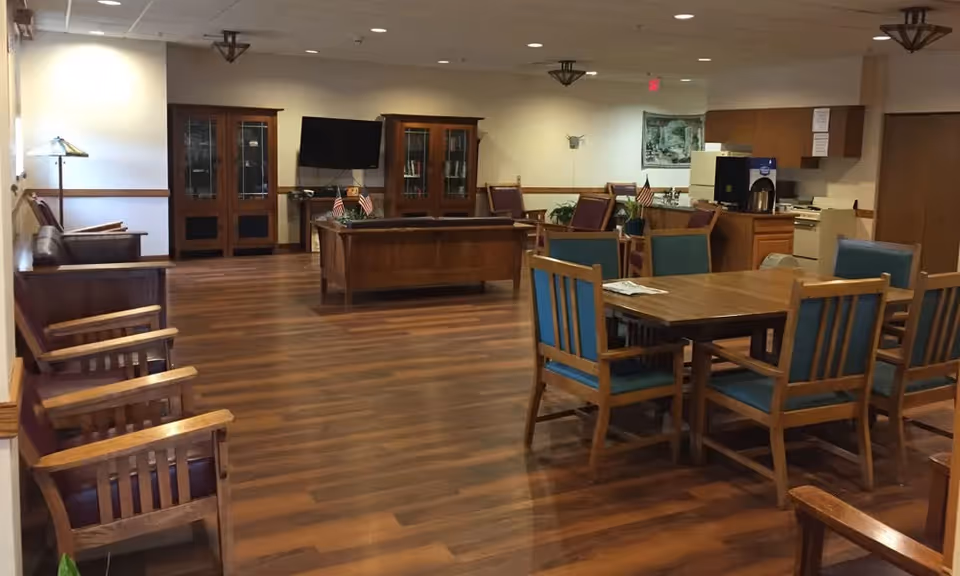 Interior view of a senior living facility common area with wooden flooring, several wooden chairs with blue cushions around a wooden table, a few armchairs along the wall, a wooden desk with small American flags, a TV mounted on the wall, and a kitchenette area in the background.