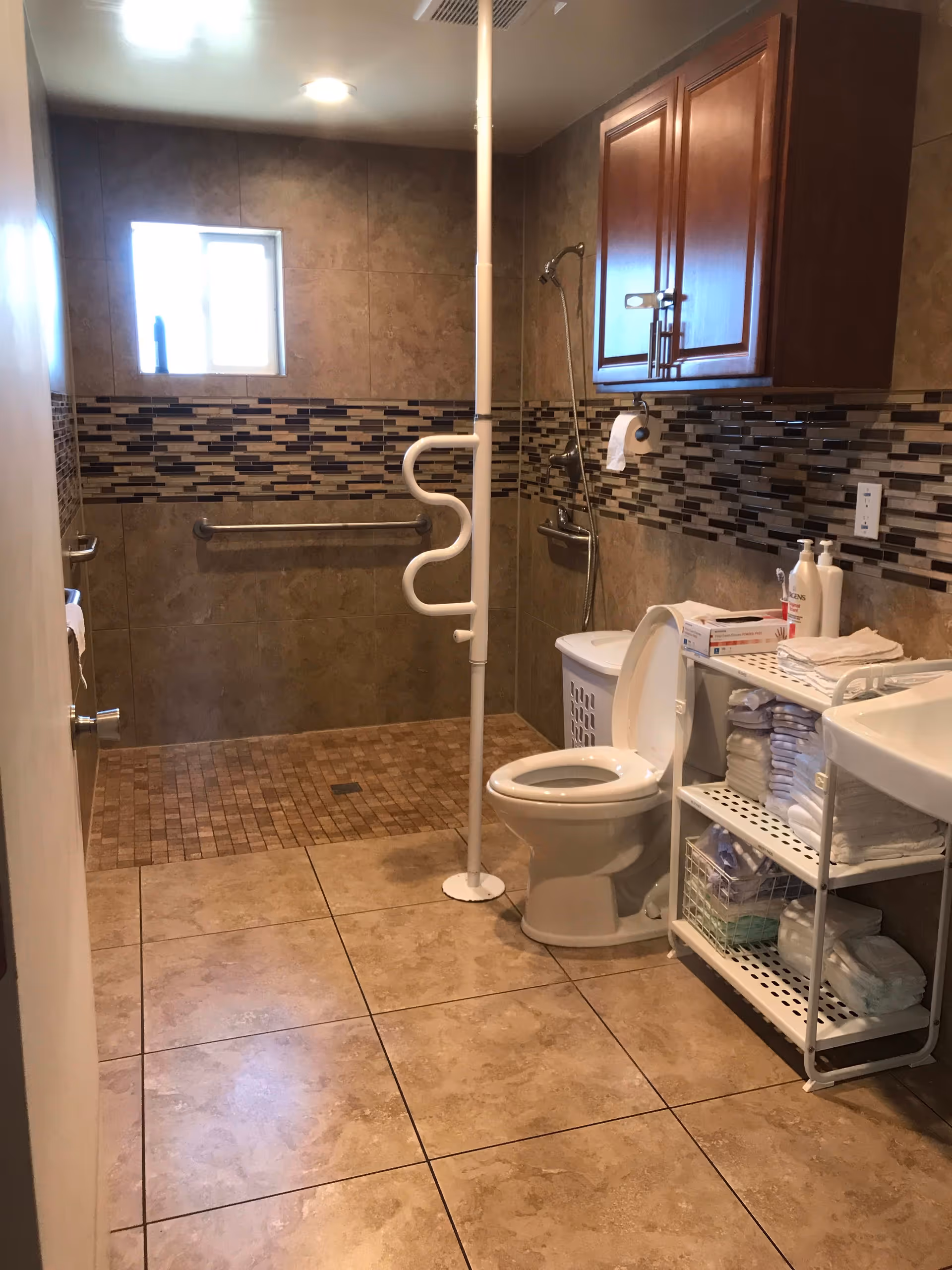 A bathroom with beige tiled floors and walls, featuring a walk-in shower area with a grab bar and a vertical support pole with a curved handle. There is a toilet with the lid open, a white shelving unit stocked with towels and toiletries, a laundry basket, and a wooden cabinet mounted on the wall above the toilet. A small window lets in natural light.