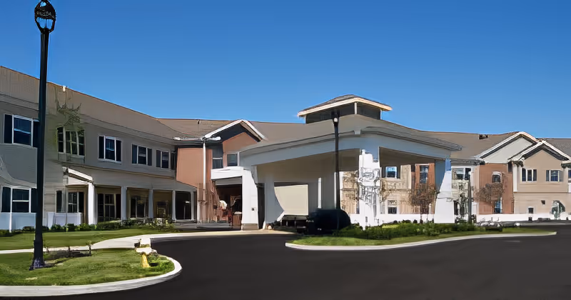 Exterior view of a senior living facility building with a covered entrance driveway, multiple windows, and a clear blue sky above.