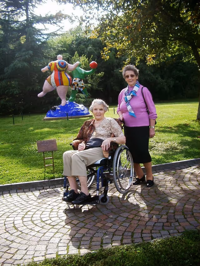 Two elderly women outdoors in a garden area; one woman is sitting in a wheelchair while the other stands beside her. Behind them is a colorful sculpture of two abstract figures playing with a red ball, surrounded by green grass and trees.