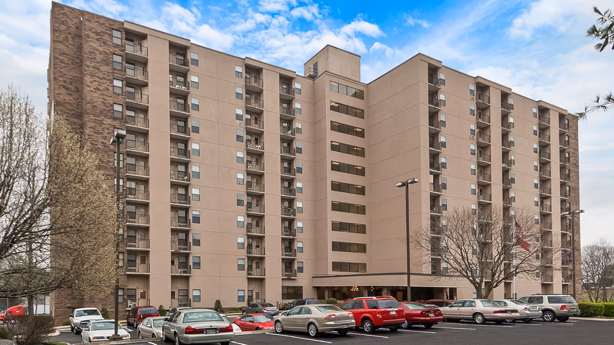 Exterior view of a multi-story senior living facility building with balconies and a parking lot in front filled with cars. The sky is partly cloudy and there are some leafless trees around the building.
