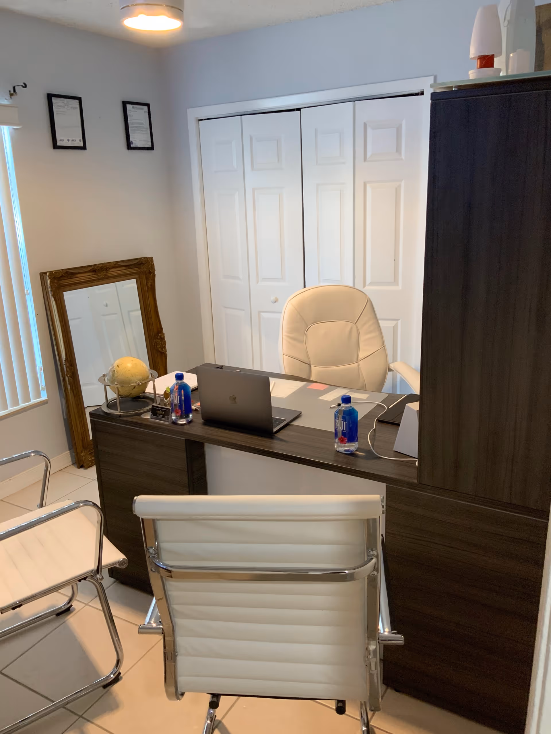 Small office with a dark wood desk, white chairs, a laptop, bottled water and a globe in front of a closet with bi-fold doors.