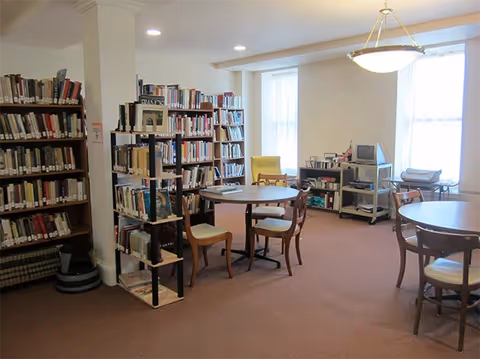 A quiet library room with bookshelves filled with books, round tables with chairs, a yellow armchair near the window, and a small TV on a stand. The room is well-lit with natural light coming through large windows.